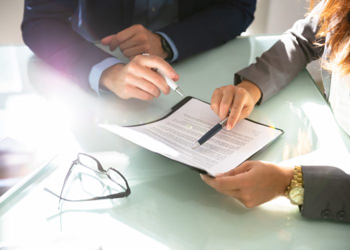 Two Businesspeople Hand Analyzing Document Over Glass Desk