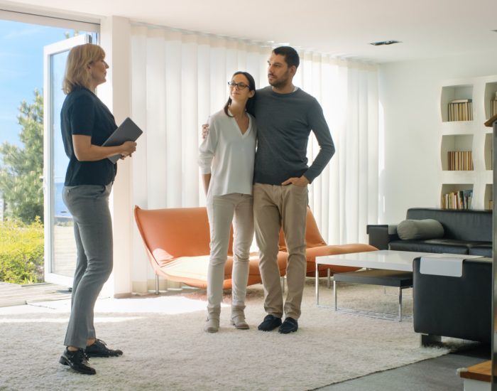 Professional Real Estate Agent Shows Stylish Modern House to a Beautiful Young Couple Who are in the Market for Purchasing/ Renting New Home. House Has Floor to Ceiling Windows and Seaside View.